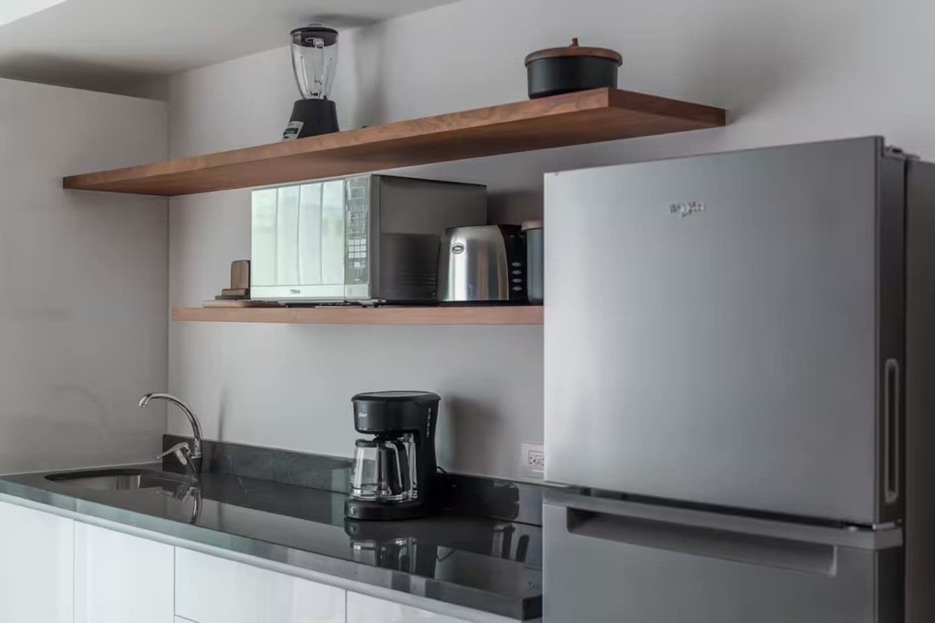 A modern kitchen with a stainless steel refrigerator, a coffee maker, a microwave, and a blender on a wooden shelf.
