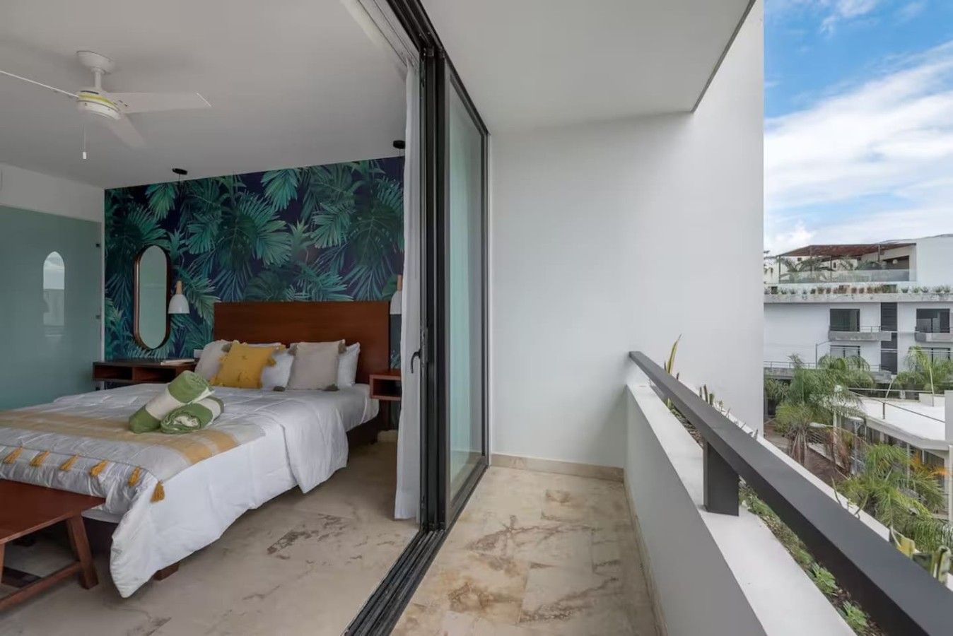 A hotel room with a bed, a nightstand, a mirror, and a view of the outside.