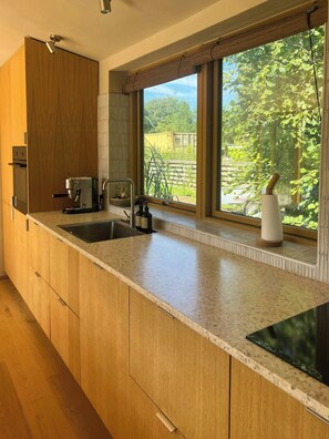 A modern kitchen with wooden cabinets, a built-in oven, a coffee machine, a sink, and a window with a view of greenery. - Sustainable house in wild garden near Odsherred (Højby)