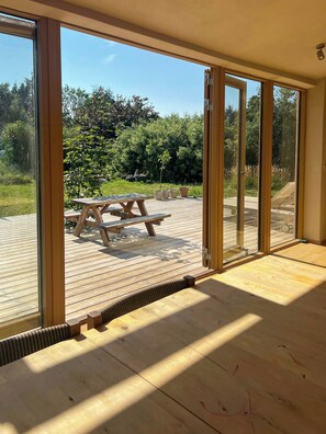 A wooden deck with a picnic table, large glass doors leading to a garden, and a clear view of the outdoors. - Sustainable house in wild garden near Odsherred (Højby)