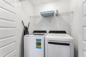 A laundry room with two front-loading washers and dryers, a metal shelf above, and a door on the left. - 🐾 Lost Bay Townhome 4239 | Dog Friendly | New Build + Heart of Orange Beach (Orange Beach)