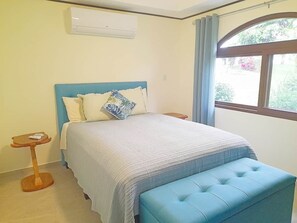 A bedroom with a bed, a bedside table, a window with curtains, and an air conditioning unit. - Villa and Casita (Tronadora)