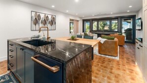 A modern kitchen with a central island, a dining area with a sofa, and a large window providing a view of the outdoors. - Luxury Condo E1 + Hot tub at Wapato Point Chelan (Manson)