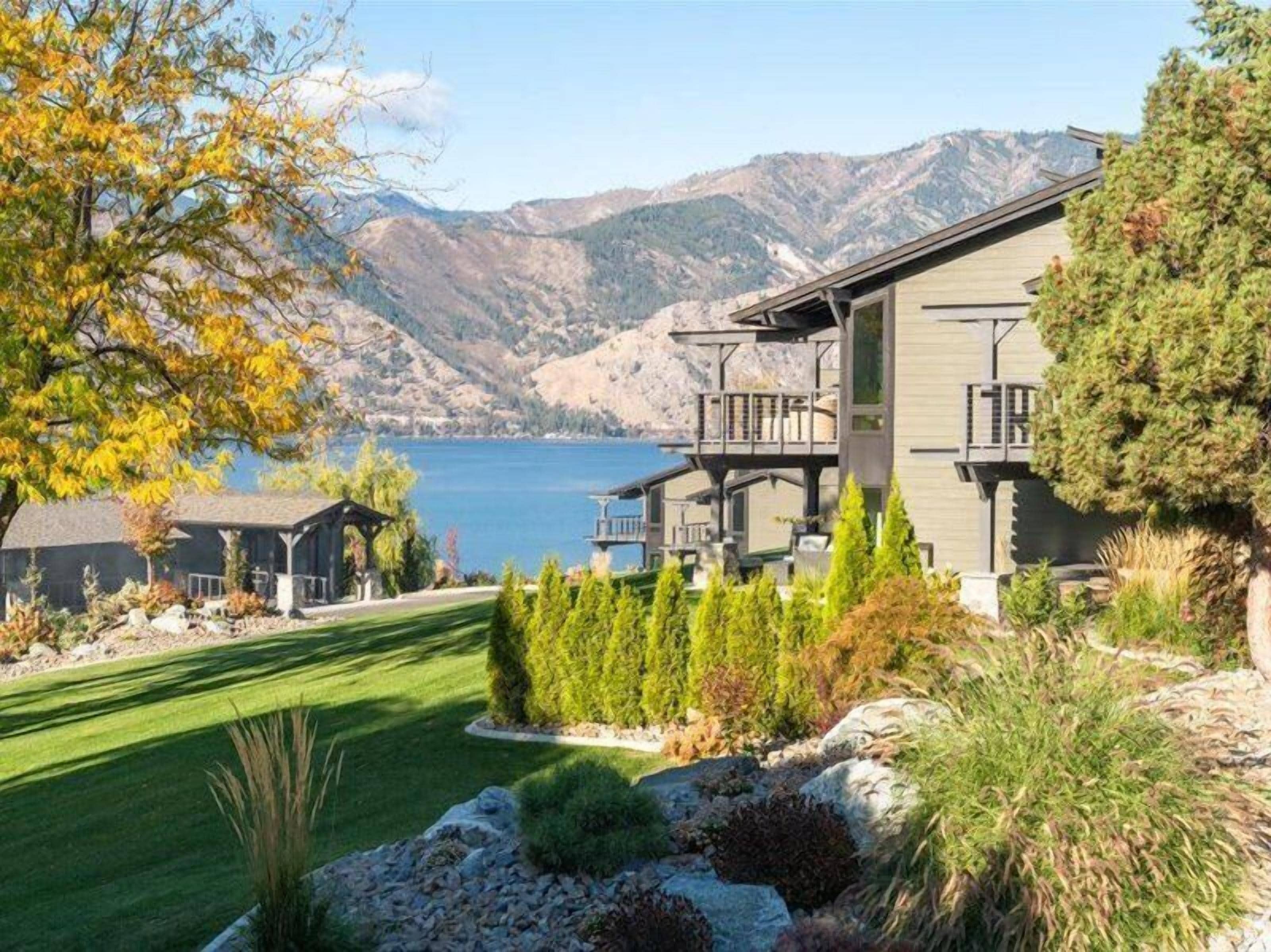 A modern two-story house with a deck overlooking a lake, surrounded by a well-maintained lawn and various shrubs.