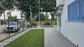 A paved deck with a neatly trimmed lawn, a white car parked in a fenced area, and a building with blue shutters.