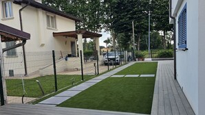 A paved deck with a wooden floor, a fenced yard with artificial grass, a white building with blue shutters, and a parked vehicle in the background. - A welcoming and spacious apartment of approximately 130 sqm—perfect for 8 guests—located within a villa in an exclusive neighborhood of Marina di Pietrasanta, bordering Forte dei Marmi.Its strategic location allows you to reach the sea in just 7 minu (Marina di Pietrasanta)
