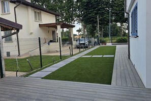 En stenlagd uteplats med trägolv, en inhägnad trädgård med konstgräs, en vit byggnad med blåa fönsterluckor och ett parkerat fordon i bakgrunden.