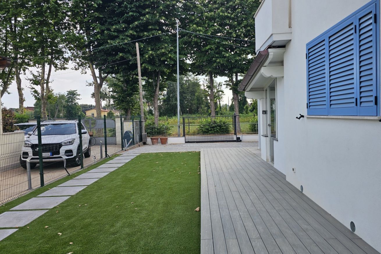 A paved deck with a neatly trimmed lawn, a white car parked in a fenced area, and a building with blue shutters.