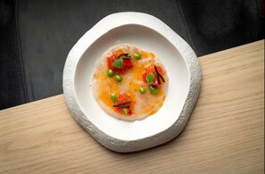 Un tazón blanco con borde texturizado que contiene un plato con guisantes, tomates y hierbas, sobre una superficie de madera.
