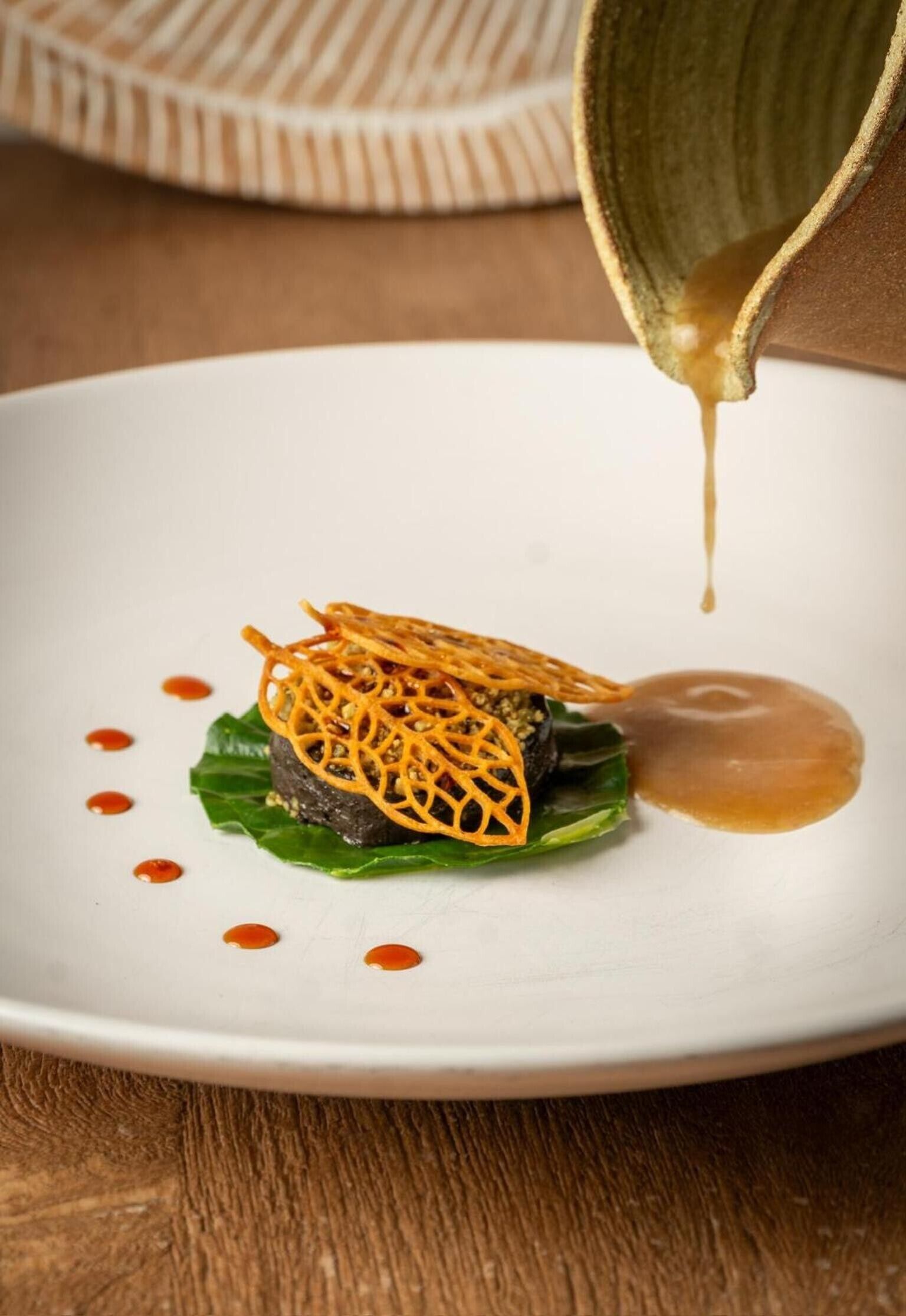 Un piatto bianco con un condimento, salsa in corso di versamento e un sottopentola decorato sullo sfondo.