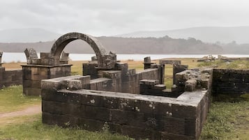 Una antigua estructura de piedra con un arco circular, rodeada de terreno cubierto de hierba y una masa de agua al fondo.