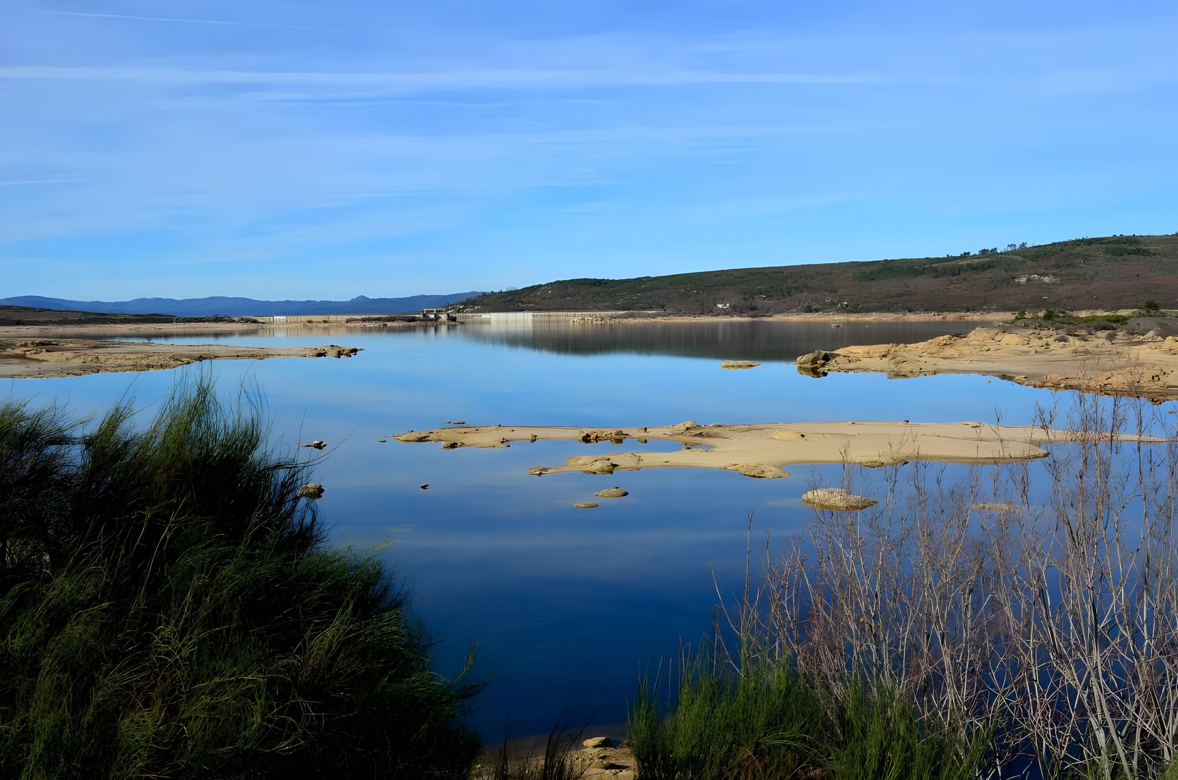 A calm lake with clear skies, surrounded by hills and sparse vegetation.