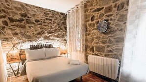 A bedroom with a stone wall, a bed, a bedside table, a radiator, and a window with curtains. - Esmeralda Double Room with Interior Patio and Wi-Fi (Porqueirós)