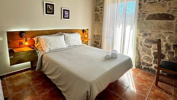 A bedroom with a bed, a nightstand, a chair, stone walls, and a window with curtains. - Zafiro Double Room with Mountain Views, Private Balcony, and Wi-Fi (Porqueirós)