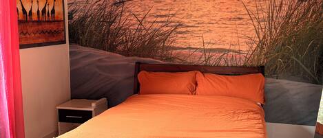 A bedroom with a bed, a nightstand, a wall-mounted lamp, and a sunset mural.