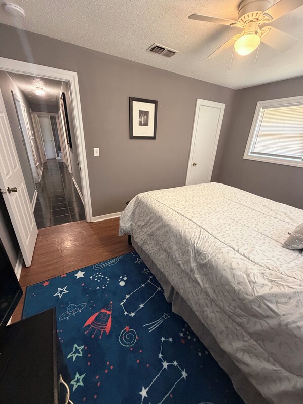 A bedroom with a bed, a ceiling fan, a window with blinds, a door, and a hallway leading to another room.