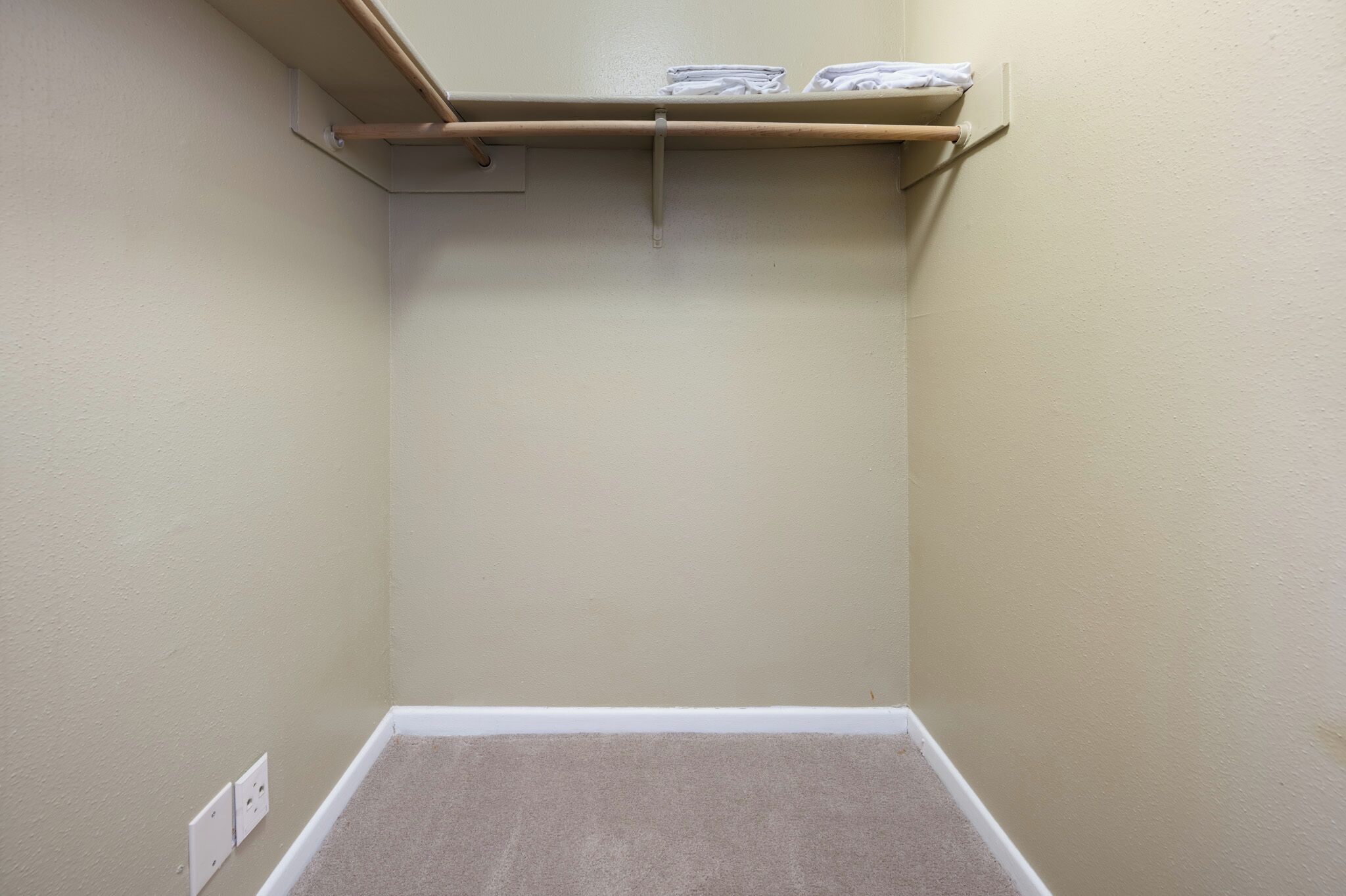 A corner room with a carpeted floor, a wooden shelf with folded towels, and a light switch on the wall.