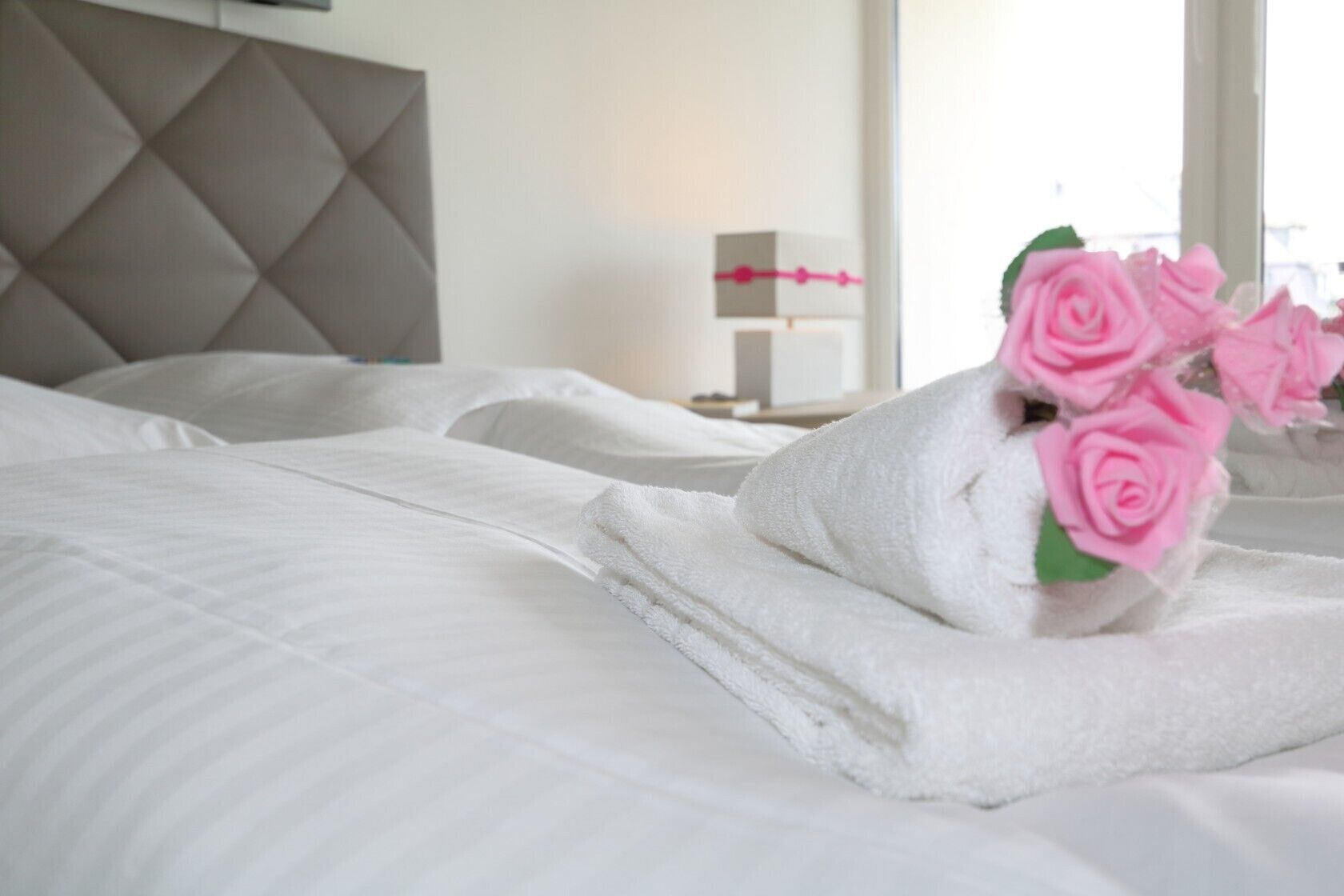 A neatly made bed with white linens and a bouquet of pink roses.