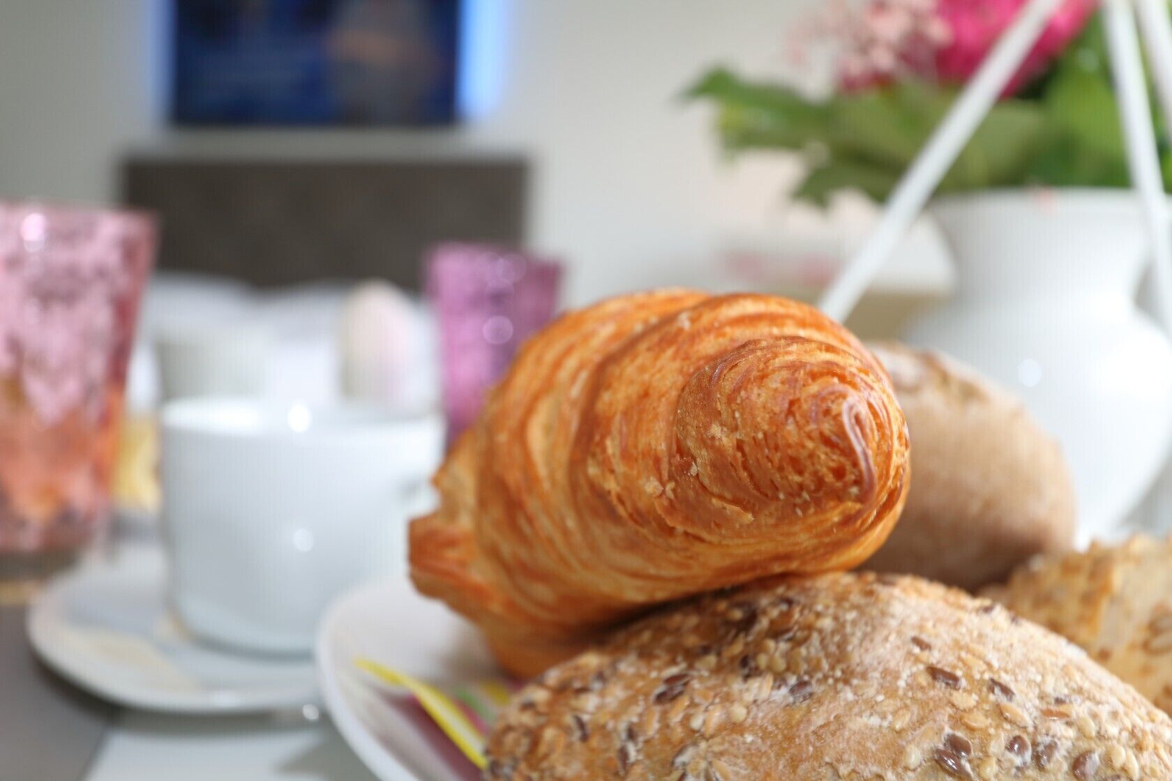 Ein Croissant und ein Brot liegen auf einem Tisch, im Hintergrund stehen Tassen und eine Blumenvase.