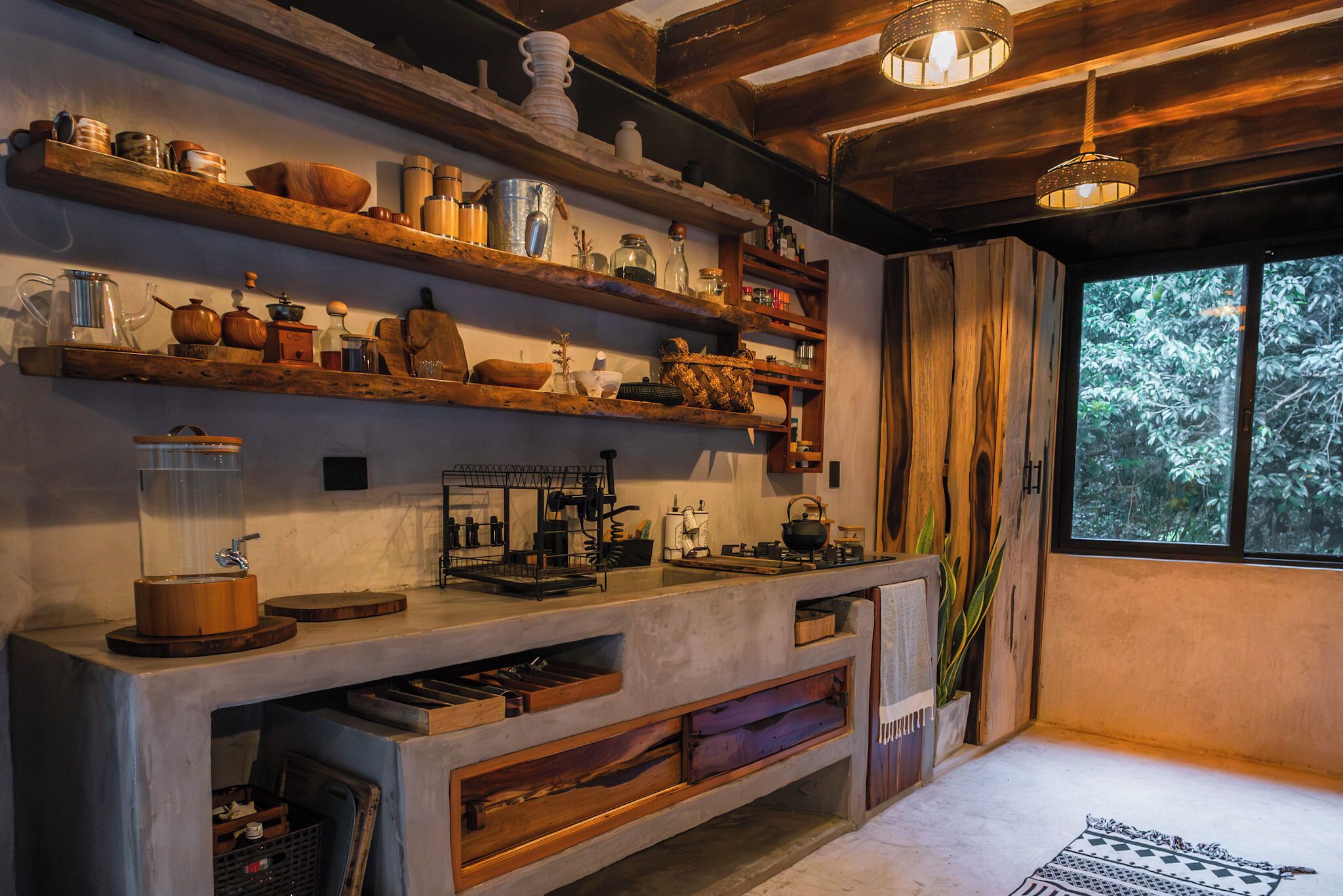 Cocina rústica con repisas de madera, encimera de concreto y ventana con vista a la vegetación.