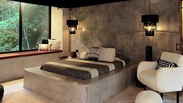 A modern bedroom with a large bed, a white armchair, and a view of greenery through the window.