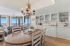 A dining area with a round wooden table, white chairs, a chandelier, and a view of the ocean through large windows. - 🏊🥽🌴Ebb Tide 406 | NSB's Finest | New Pool + Sundeck + Beach Access (New Smyrna Beach)