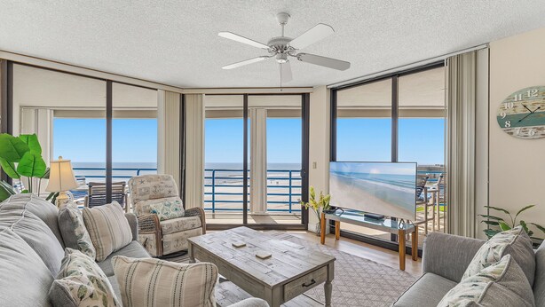 A spacious living room with a large TV, comfortable seating, a ceiling fan, and a view of the ocean. -    🏊🥽🌴Ebb Tide 406 | NSB's Finest | New Pool + Sundeck + Beach Access (New Smyrna Beach)