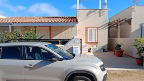 En sølvgrå SUV parkeret foran en bygning med et tagtækket tag, en parabolantenne og krukkeplanter.