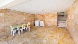Une pièce avec un sol carrelé, une table entourée de chaises, un petit meuble blanc et un escalier menant à l’étage supérieur.