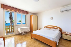 Ein Schlafzimmer mit Bett, Nachttisch, Kleiderschrank, Tisch mit Tischdecke und Blick auf das Meer durch eine Schiebetür.