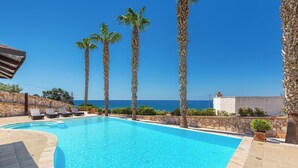 Une piscine à l’eau d’un bleu cristallin, entourée de murs de pierre et de palmiers, avec une vue sur la mer en arrière-plan.
