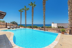 A swimming pool with clear blue water, surrounded by stone walls and palm trees, with a view of the sea in the background.