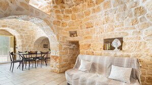A stone-walled room with a sofa, dining table, chairs, and a small shelf with decorative items. - Trullo Sobba Llera by BarbarHouse (San Michele Salentino)