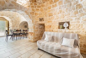 Una habitación con paredes de piedra, un sofá, una mesa de comedor, sillas y un pequeño estante con objetos decorativos.