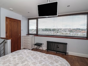 Ein Schlafzimmer mit Bett, Kommode, Heizkörper, Fernseher, Fenster mit Jalousien und einer Tür.