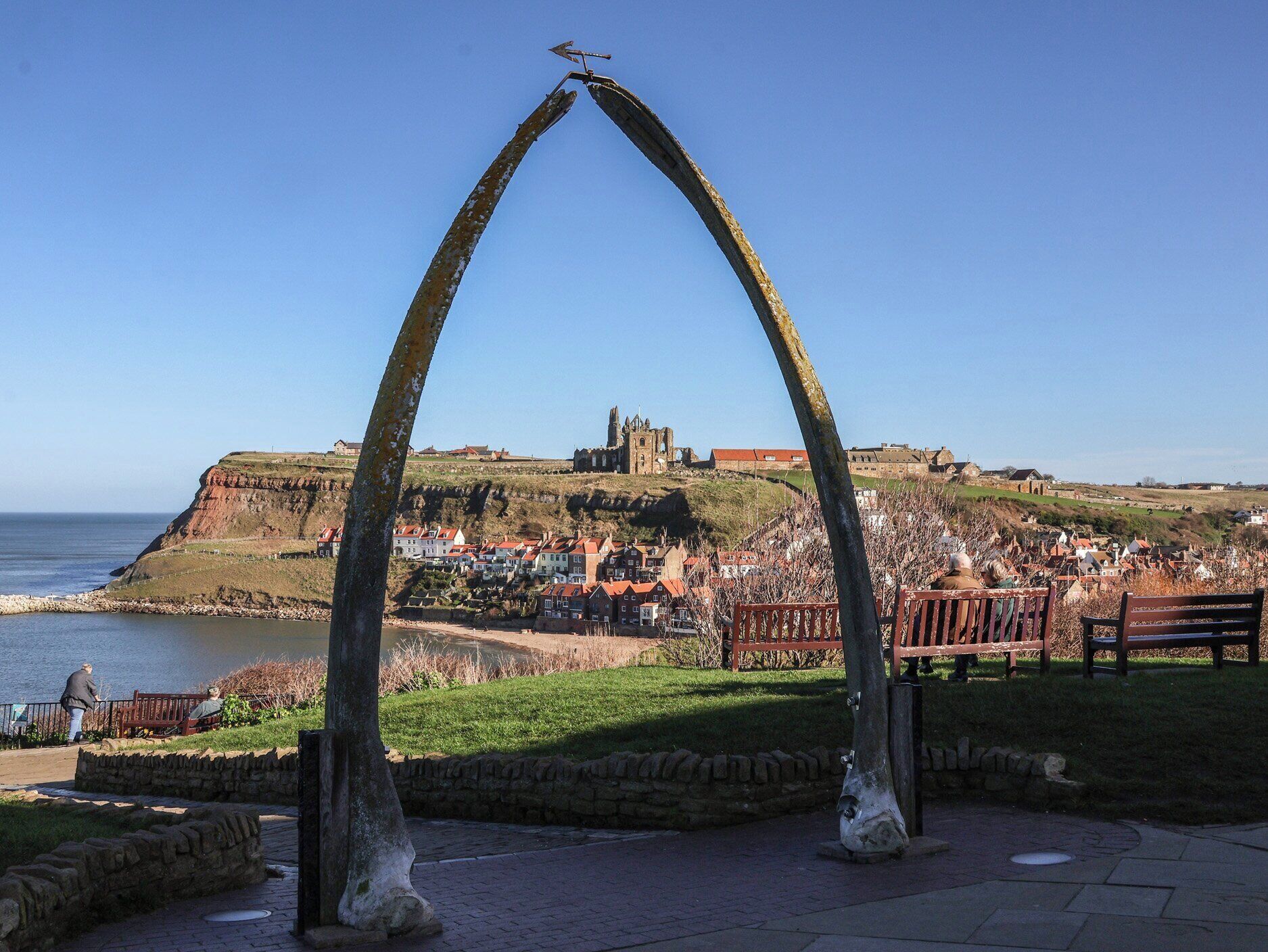 A park with a metal archway, benches, and a view of a historic building across the water.