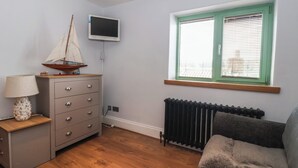 Um quarto com piso de madeira, penteadeira cinza, luminária, um modelo de veleiro, televisão na parede e janela com persianas.