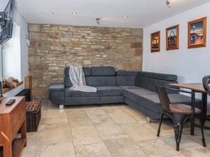 A modern living room with a stone wall, grey sectional sofa, a small dining table with chairs, and a TV mounted on the wall. - 5 Hall Yard (Whitby)