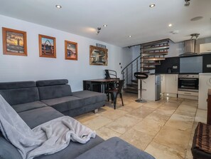 Ein modernes Wohnzimmer mit grauem Sofa, einer Essecke mit Tisch und Stühlen, einer Küche mit Geräten und einer Treppe.