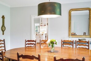 A dining room with a round wooden table, six wooden chairs, a vase with flowers, a large green pendant light, a wall clock, and a mirror with a gold frame. - Shelter Island Estate · Heated Pool · Guest Cottage · Walk to Beach · Sleeps 10 (Shelter Island Heights)