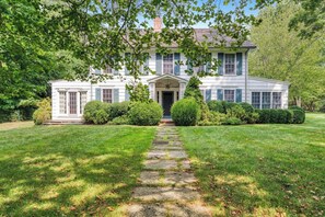 A two-story house with a central entrance, surrounded by a well-maintained lawn and trimmed hedges. - Shelter Island Estate · Heated Pool · Guest Cottage · Walk to Beach · Sleeps 10 (Shelter Island Heights)