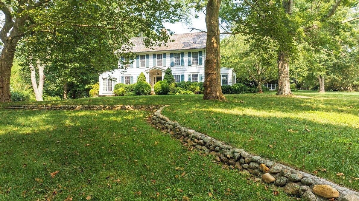 Une grande maison avec un chemin de pierre menant à l’entrée, entourée d’une végétation luxuriante et d’arbres.
