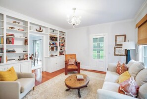 A well-lit living room with a fireplace, a bookshelf, a round coffee table, and a comfortable seating area. - Shelter Island Estate · Heated Pool · Guest Cottage · Walk to Beach · Sleeps 10 (Shelter Island Heights)