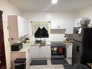 A compact kitchen with white cabinets, a black stove, a refrigerator, and a window with a curtain. - Savannah Sunset- Charming 2 Bedroom Home with AC and Community Pool (Runaway Bay)