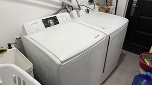 A laundry area with two white washing machines, a white laundry basket, and a blue water container. - Savannah Sunset- Charming 2 Bedroom Home with AC and Community Pool (Runaway Bay)