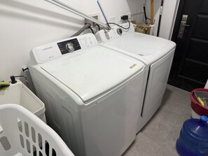 A laundry area with two white washing machines, a white laundry basket, and a blue water container. - Savannah Sunset- Charming 2 Bedroom Home with AC and Community Pool (Runaway Bay)