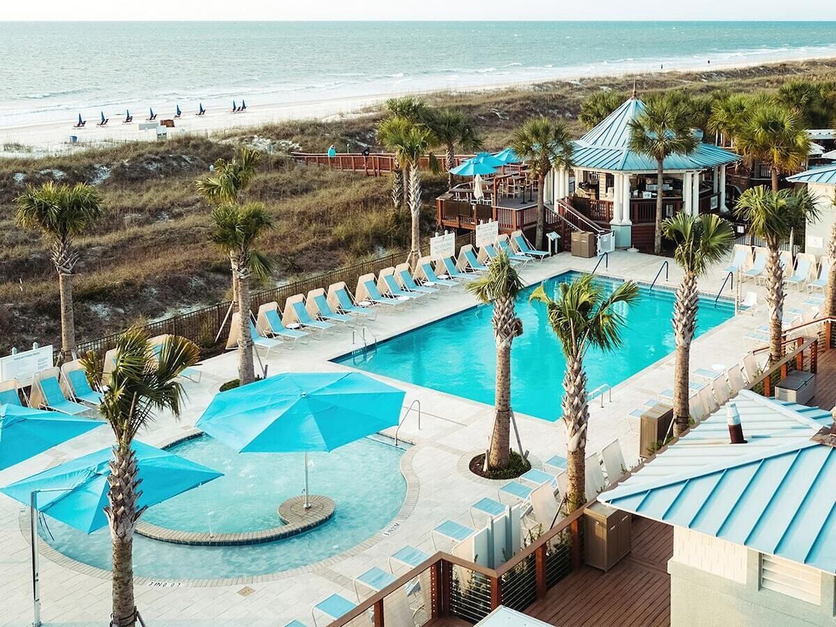 Un espace piscine avec des chaises longues et des parasols, une vue sur la plage et un bâtiment avec un toit bleu.
