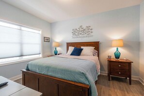 A bedroom with a bed, bedside tables, a lamp, a wall decoration, and a window. - Breezy Beach Haven - 3rd story 1 bedroom condo w ac, laundry, deck, elevator. (Hampton)