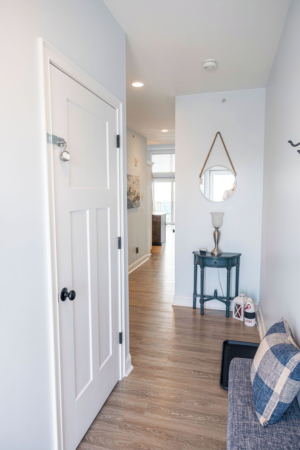 A hallway with a white door, a blue table with a lamp, a mirror, and a checkered pillow on a couch.