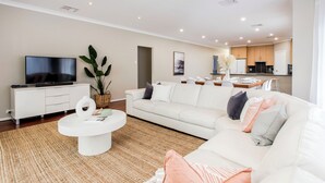 Una sala de estar moderna con un televisor de pantalla plana, sofás blancos, una mesa de centro y una cocina al fondo.